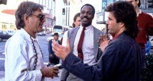 Three men talk on a city street: one in sunglasses and holding headphones, one in a suit, and one gesturing in a dark jacket. Buildings behind.