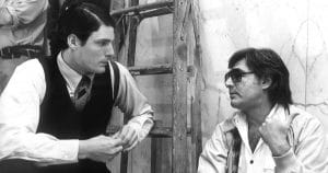 Two men sit and talk by a wooden ladder; one in a vest and tie, the other in sunglasses with an open shirt over a t-shirt, both serious.