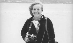 A smiling woman stands outdoors by the water, holding a Nikon camera and wearing a camera strap around her neck. She has shoulder-length hair and is dressed in a dark jacket.