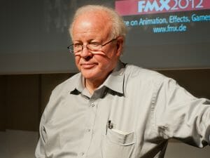 An older man with white hair and glasses stands indoors in a light gray shirt with a pen in his pocket. A screen shows FMX 2012 info.