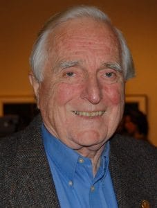 An elderly man with white hair smiles while wearing a blue collared shirt and a gray blazer, standing indoors with blurred people and artwork in the background.