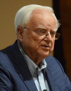 An elderly man with white hair and glasses, in a blue blazer and light blue shirt, speaking at a meeting.