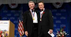 Two performers on stage in tuxedos, each with gold medals worn around their necks on red, white, and blue ribbons. One is a generation older than the other.