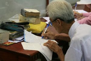 An elderly person with gray hair writes on sheets of paper at a cluttered desk; another person writes in the background.