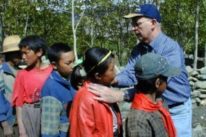 A doctor in a blue cap and glasses checking the necks of children lined up outdoors, with trees and rocks behind them.