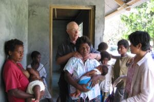 A group stands outside a building; two women hold babies, with a man behind her, checking one woman's neck.