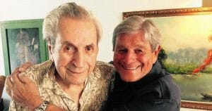 Two older men smile and embrace indoors; one wears a patterned shirt, the other a dark one. A colorful landscape painting is behind.
