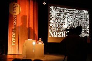 A speaker stands at a lectern onstage at TYPO Berlin 2008 with a large screen of white font names. A cameraman films from the side.