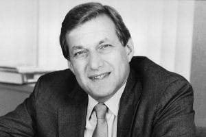 A black and white photo of a middle-aged man in a suit and tie, smiling at his desk with bookshelves behind him.