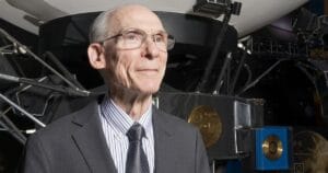An older man in a suit and tie stands before a large spacecraft model with gold discs at a space museum exhibit.
