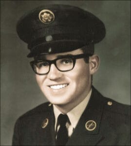 A young man in U.S. Army Vietnam-era dress uniform and glasses smiles at the camera. He wears a peaked cap and jacket adorned with military insignia, set against a plain studio background.