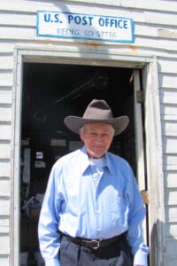 An older man in a cowboy hat and blue shirt smiles in the doorway of a small Redig, SD Post Office as sunlight falls on him.