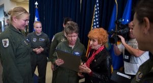 A group of women in military uniforms stand together as an older woman is presented a framed document; flags and a cameraman behind.