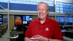 A smiling older man in glasses and a red WKYC 3 polo shirt stands in a modern newsroom with desks, computers, and large screens.
