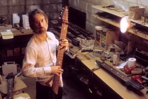 A bearded man with glasses holds a large stringed instrument in a cluttered workshop lit by a bright desk lamp.