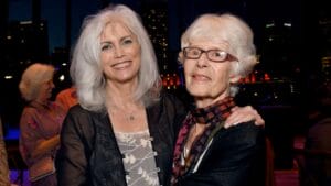 Two older women stand close together, smiling indoors at night. City lights and guests behind them; one wears glasses and a scarf.