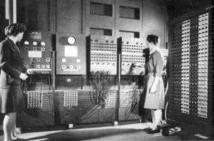 Two women operate a large, early computer with numerous cables, switches, and dials. They stand in front of the machine, which fills the room and features panels covered in wires and connectors.