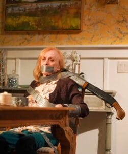 A close-up of a film clip showing an older woman, tied to a chair with duct tape at a dining table, looks distressed in a warmly lit room with floral wallpaper.