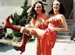 Two women dressed as Wonder Woman, one holding the other in her arms. They smile and pose outside near a building and stairs.
