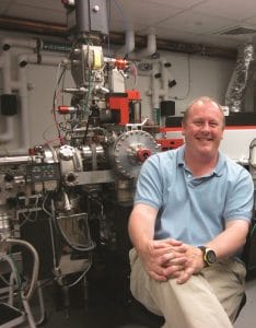A man in a light blue polo shirt sits smiling with arms crossed in front of a large scientific machine with pipes, wires, and metal components in a laboratory setting.