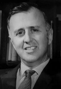 A man in a suit and tie poses for a formal portrait. It is black and white.