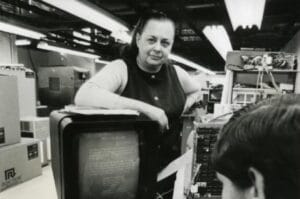 A woman stands in a computer lab, resting her arm on a vintage computer monitor displaying text, with equipment and electronic components visible in the background.