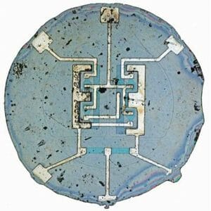 Close-up of an integrated circuit chip with detailed metallic connections on a round, slightly worn blue-gray silicon wafer.