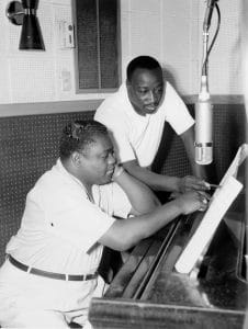 Two men in a recording studio examine sheet music atop a piano. One man sits at the piano, while the other leans over beside him, both focused on the music. A large microphone hangs above them. The photo is in black & white.