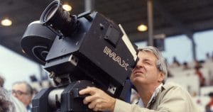 A man uses a large IMAX film camera outdoors, peering through the viewfinder, with blurred spectators and seating behind.