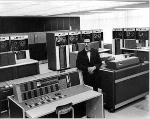 A man in a suit and bow tie stands in a large room with vintage mainframe computers, reel-to-reel tape drives, and control panels from the mid-20th century.