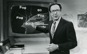 A male weather presenter in a suit points at a screen weather map showing “Fog,” 60°F, “Tonight,” and “Cloudy” in various areas.