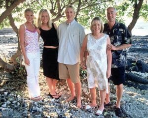 Five people stand on a beach, smiling at the camera. Three women and two men wear summer clothes; trees and rocks behind.