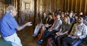 An elderly man stands in front of a group of college-age students, who are paying close attention to him.