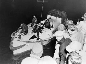 A group of people watch as three men in dark clothing stand on a small lifeboat at night, with ropes securing the boat and equipment visible on deck.