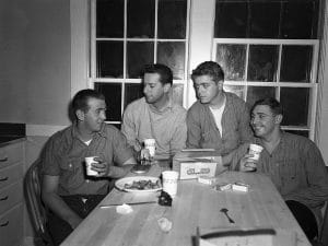 Four young men sit around a kitchen table, smiling and holding cups. There is a plate of food, a box, and assorted items on the table. Large windows are behind them, and they appear to be enjoying a casual gathering.