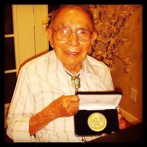 An elderly man wearing glasses and a bolo tie smiles while holding an open case displaying a large gold medal. He is seated indoors with a flowering plant and a door in the background.