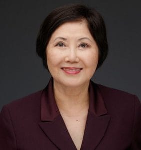 A woman with short dark hair in a burgundy blazer smiles at the camera, standing against a dark gray background.