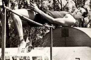 A male athlete arches over a high jump bar, legs and arms bent. He wears sports gear. Trees and a building in the black-and-white background.