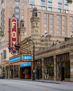Street view of the historic Fox Theatre in Atlanta, featuring its distinctive marquee, tall neon sign, ornate architecture, and a modern building in the background.