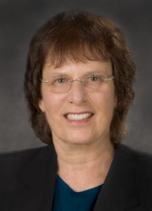 A middle-aged woman with short brown hair and glasses smiles at the camera. She is wearing a dark blazer over a teal top, with a neutral gray background.