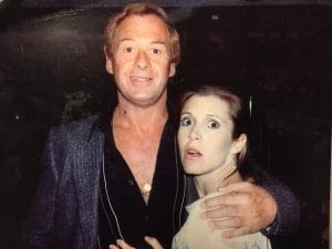 A man with light hair smiles and gently holds a surprised-looking Carrie Fisher. They are standing close together in a dimly lit setting.