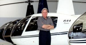 An older man with glasses smiles with arms crossed in front of a white R44 Raven helicopter inside a hangar, others behind him.