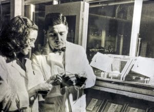 Two scientists in lab coats examine an object together in a mid-20th-century lab, standing by cages and scientific equipment.