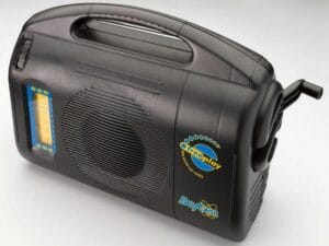 A black portable radio with a large speaker, a yellow and blue tuning scale, a handle on top, and a hand crank on the side. The brand Freeplay is labeled on the front.