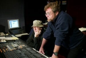 Two men in a recording studio focus on an audio mixing console; one sits with a bucket hat and glasses, the other stands nearby.