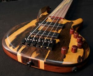 A close-up of an electric bass guitar with light and dark wood body, six tuning knobs, metal bridge, and pickups on black.