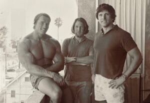 Three men pose together on a balcony; one is shirtless and muscular, while the other two are wearing casual shirts. The background features palm trees and buildings, indicating a warm, coastal environment.