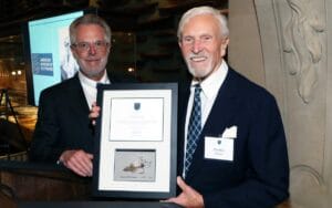 Two men in suits stand indoors; one hands a framed certificate and drawing to the other, who smiles at the camera. In the background, there is a museum display and a screen displaying the words "American Museum of Fly Fishing."