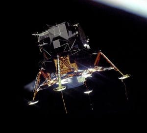The Apollo 11 lunar module with gold and silver foil descends in space against a dark background, with just the edge of the moon showing in the upper right corner.