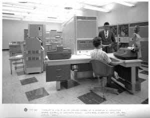 Three people—two women and one man—operate vintage computer equipment in a 1960s office with a mainframe, with a desk and typewriter-style terminal.